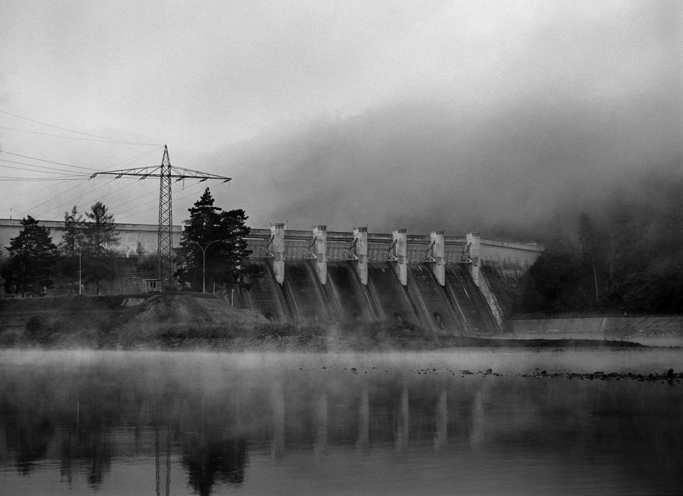 Black and white picture of a dam over tranquil water, in the background there is a mountain or a hill shrouded in fog.