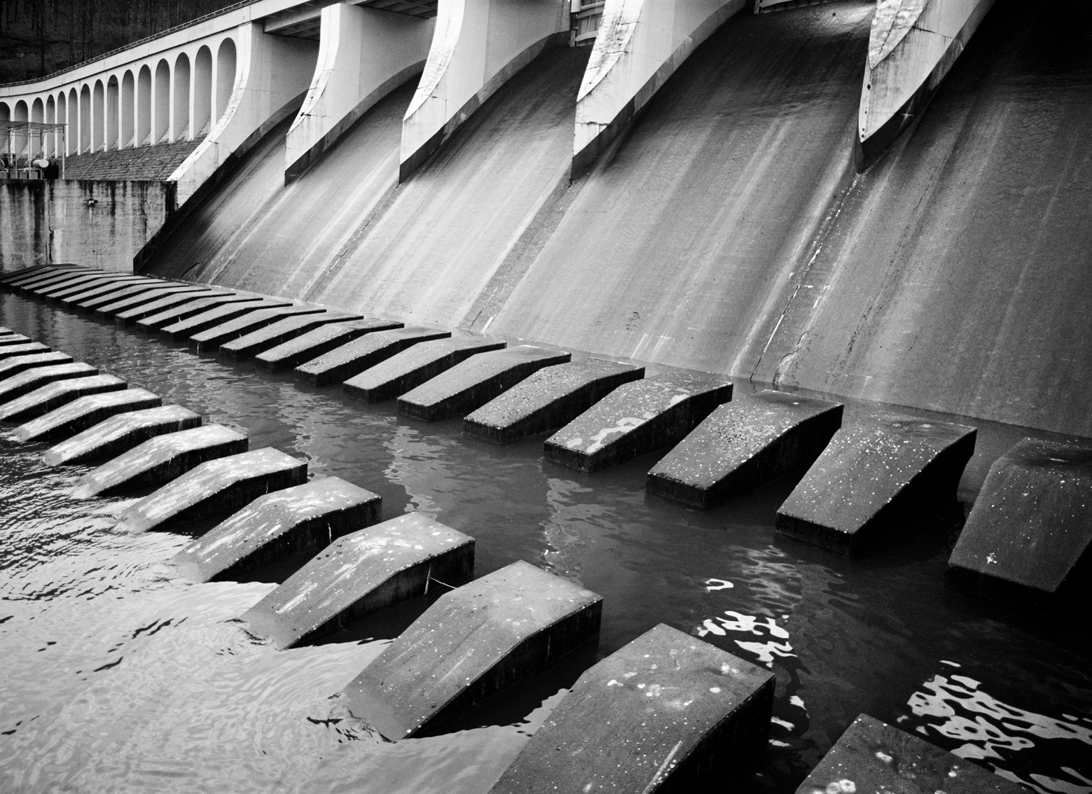 Black-and-white photograph of a concrete dam with multiple spillways and rows of concrete barriers in the water below.
